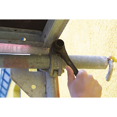 Hand mit schwarzem Werkzeug an einem Gerüst. Metallkonstruktion, gelbe Wand im Hintergrund.